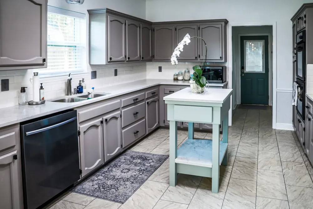 A renovated kitchen with painted gray cabinets, marble countertops and a small portable island